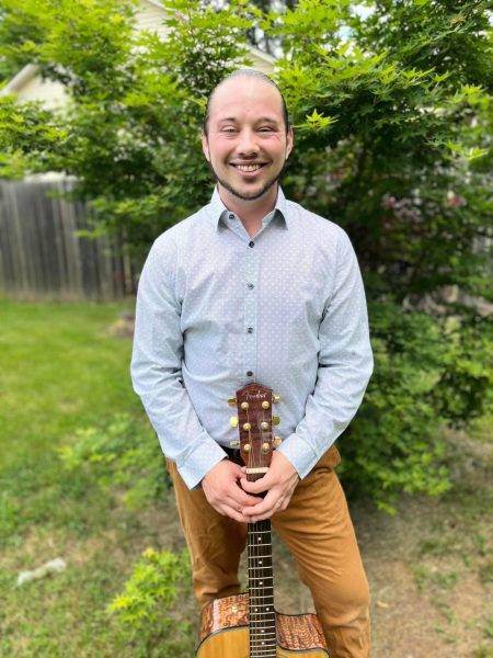 A smiling person stands outdoors holding an acoustic guitar vertically. They are wearing a light blue shirt and tan pants. A wooden fence and green foliage are visible in the background.