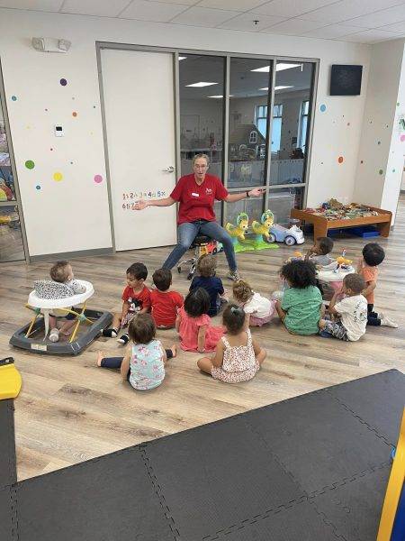 A teacher wearing a red shirt sits on a chair in front of a group of young children seated on the floor in a classroom. The children are facing the teacher, engaged in what appears to be a lesson or activity.