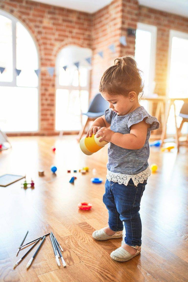 A toddler stands on a wooden floor, playing with a yellow cup. Various toys and objects are scattered around. The background features a brick wall and large windows with sunlight streaming in.